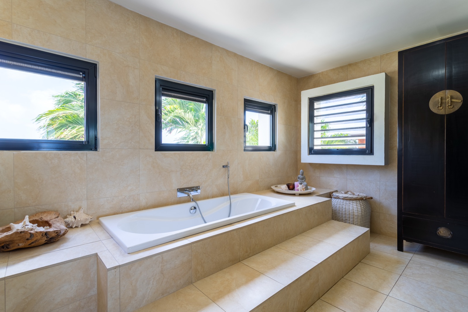 Luxury Bonaire villa spa bathroom with a deep soaking tub, natural stone tile, and windows framing tropical palm trees.